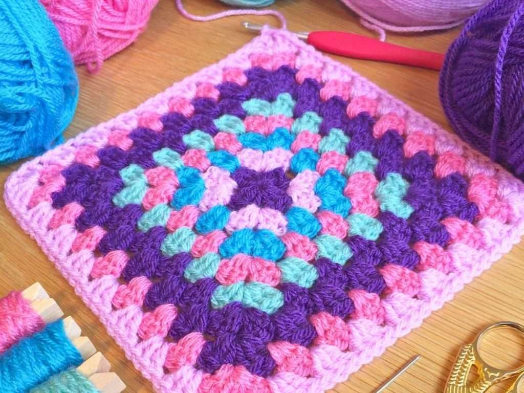 Granny Square made in blue, pink and purple with a red crochet hook and surrounded with balls of yarn
