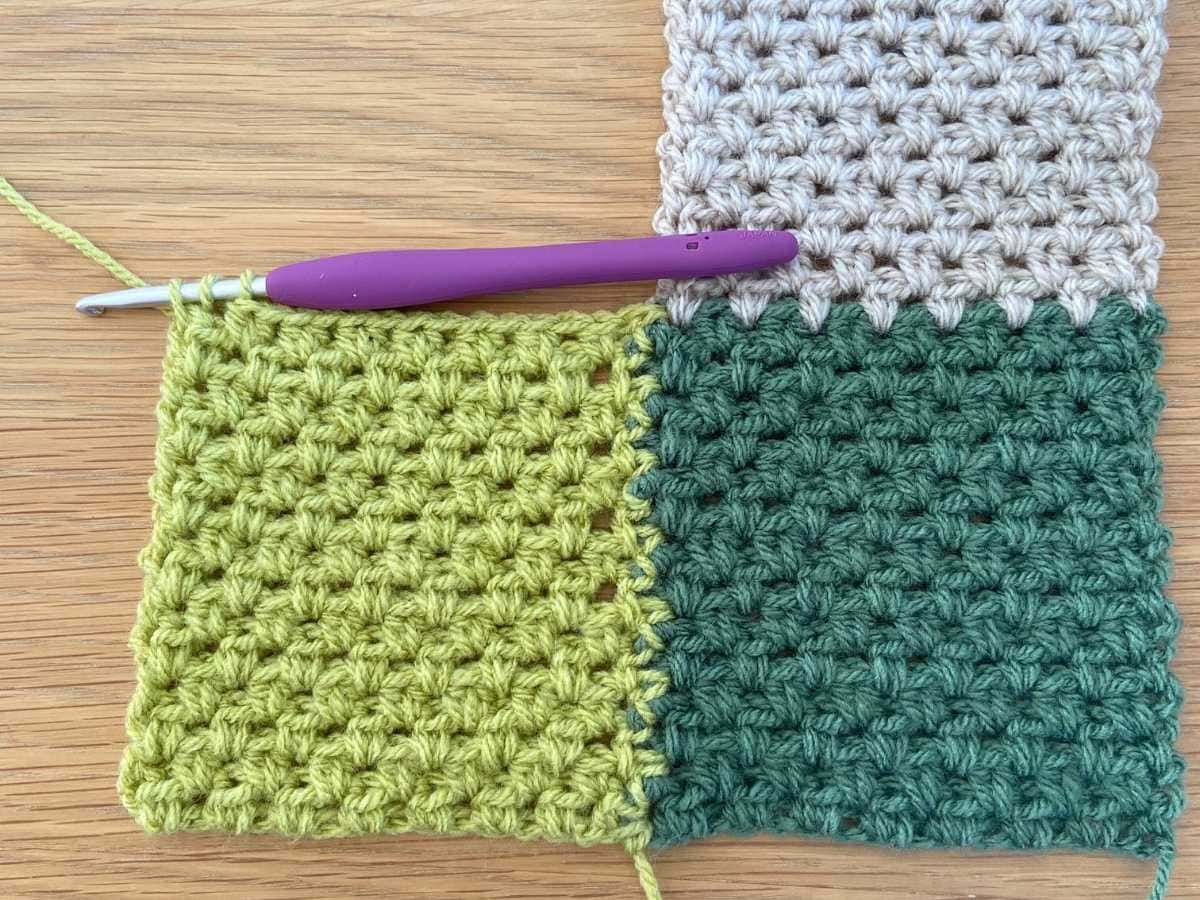 Crochet squares in green, white, and teal with crochet hook on wooden surface.